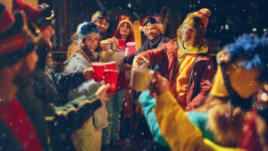 Festive Vibes Fill Air As Friends Dressed In Winter Clothes Toast Together Under Snowfall, Enjoying Warm Drinks And Cheerful Company.
